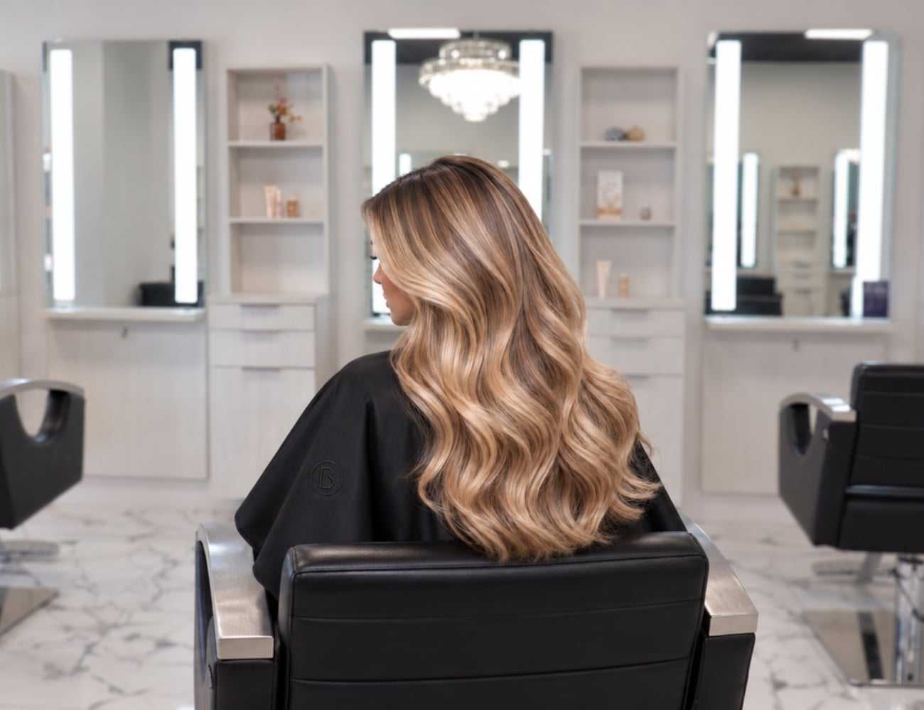 Woman with long, wavy blonde hair in salon chair, modern salon interior, mirrors on walls.